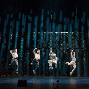  Four performers dressed in white floating over a stage with a blue curtain on the back. 