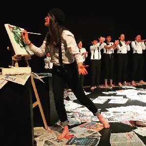  A woman artist paints over a canvas while standing on top of newspapers. A line of younger people are on the back moving their hands. 