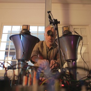 A man interacts with an electronic sound device. Cables, a microphone and two funnel-shaped columns are seen